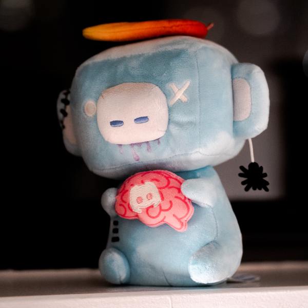 A photo of the Zombie Wumpus plushie. It’s holding a brain, has zombie-like stitches all over, and has a spider crawling from its ear. 